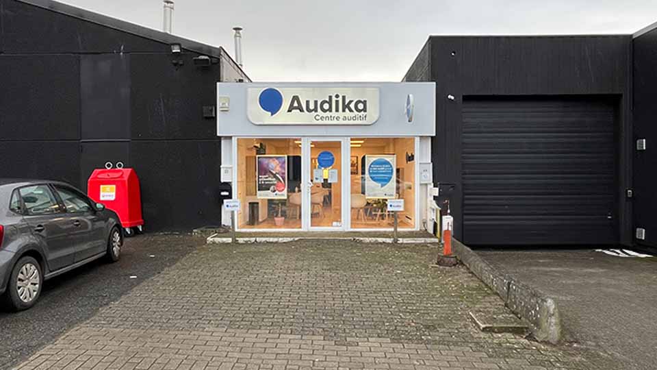 La façade d’un centre auditif Audika affiche son logo et le texte ’Centre auditif’ sur un panneau blanc. Devant, une voiture stationnée et une boîte de collecte rouge.