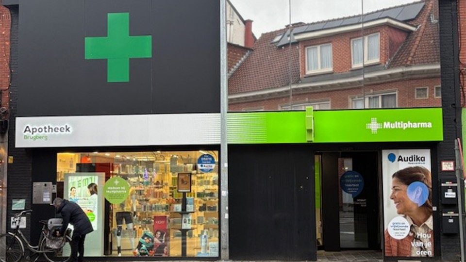 Une façade urbaine montre une pharmacie ’goed apotheek - hoorcentrum’ avec une croix verte, une entrée voisine pour un centre auditif Audika marqué par un drapeau. Contexte: magasins adjacents.