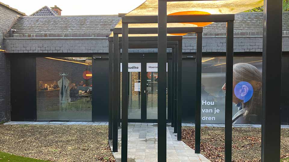 Une entrée de clinique Audika, spécialisée en soins auditifs, avec des affiches promotionnelles. Texte visible : « Hou van je oren ». Un chemin couvert mène à la porte principale vitrée.