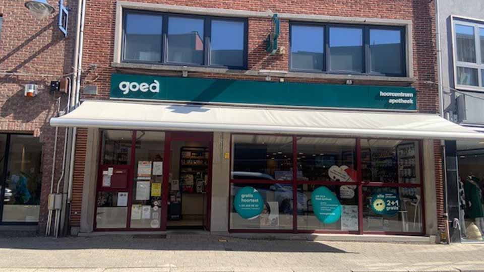 La façade d’un centre auditif/optique nommé « goed » affiche une enseigne verte avec « hoorncentrum apotheek. » Des vitrines présentent des offres : « gratis hoortest » et « 2+1 gratis. »