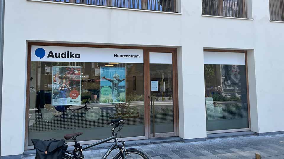 La façade d’un centre Audika ’Hoorcentrum’ présente une vitrine avec affiches publicitaires sur les soins auditifs. Un vélo est stationné devant, sur un trottoir urbain.