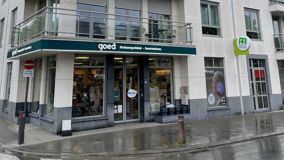 La façade d’un centre ’goed thuiszorgwinkel-hoorcentrum’ arbore une vitrine avec affiches, notamment Audika, montrant une oreille équipée d’un appareil auditif. Contexte : rue mouillée et panneaux environnants.