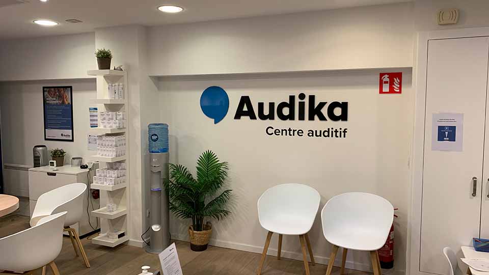 Un mur blanc affiche le logo ’Audika Centre auditif’ à côté de deux chaises blanches. L’environnement comprend une étagère avec des produits, un distributeur d’eau et des plantes décoratives, suggérant une salle d’attente accueillante dans un centre auditif dédié aux tests de l’audition et aux appareils auditifs.