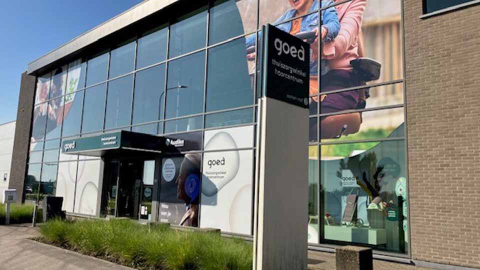 La façade d’un centre auditif ’goed,’ lié à Audika, montre des vitrines décorées avec des visuels de personnes heureuses et des inscriptions : ’Thuiszorgwinkel hoorcentrum’ et ’Hoor van je leven.’