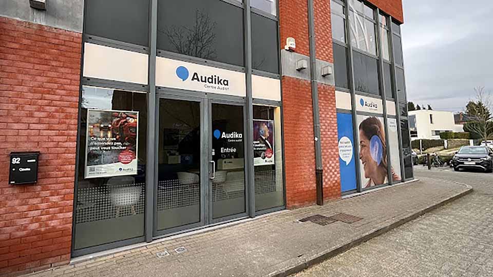 La façade d’un centre auditif Audika affiche des vitrines avec posters et textes : « Ce que vous n’entendez pas peut vous toucher », à une adresse au 92. Environnement urbain calme.
