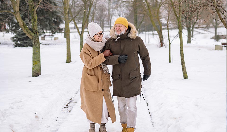 Couple marchant côte à côte sur un chemin enneigé dans un parc en hiver, habillé chaudement avec des manteaux et des accessoires d’hiver.