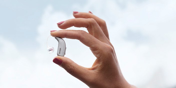 Main tenant un appareil auditif Oticon devant un ciel bleu avec des nuages.