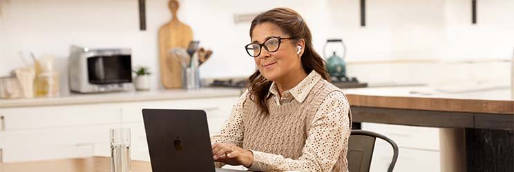 Une femme portant un appareil auditif discret travaille sur un ordinateur portable Apple dans une cuisine lumineuse moderne avec des étagères ouvertes, un micro-ondes et un verre d'eau à proximité.