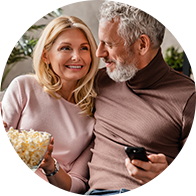 Un couple âgé partage un moment chaleureux sur un canapé, tenant du pop-corn et une télécommande.