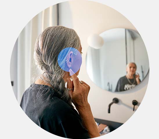 Une femme aux cheveux gris ajuste son appareil auditif devant un miroir dans une salle de bain moderne éclairée, reflétant une atmosphère calme et intime. Aucun texte visible.