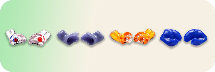 Une série de six embouts auditifs colorés (transparents, violets, orange et bleus) alignés sur un fond dégradé vert à beige, utilisés pour des appareils auditifs personnalisés.