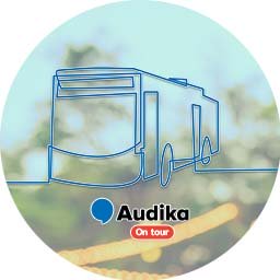 Un bus stylisé en lignes bleues semble stationné, sur un fond flou évoquant des arbres et des lumières. Texte inscrit : « Audika On tour ». Contextualisé pour la santé auditive.
