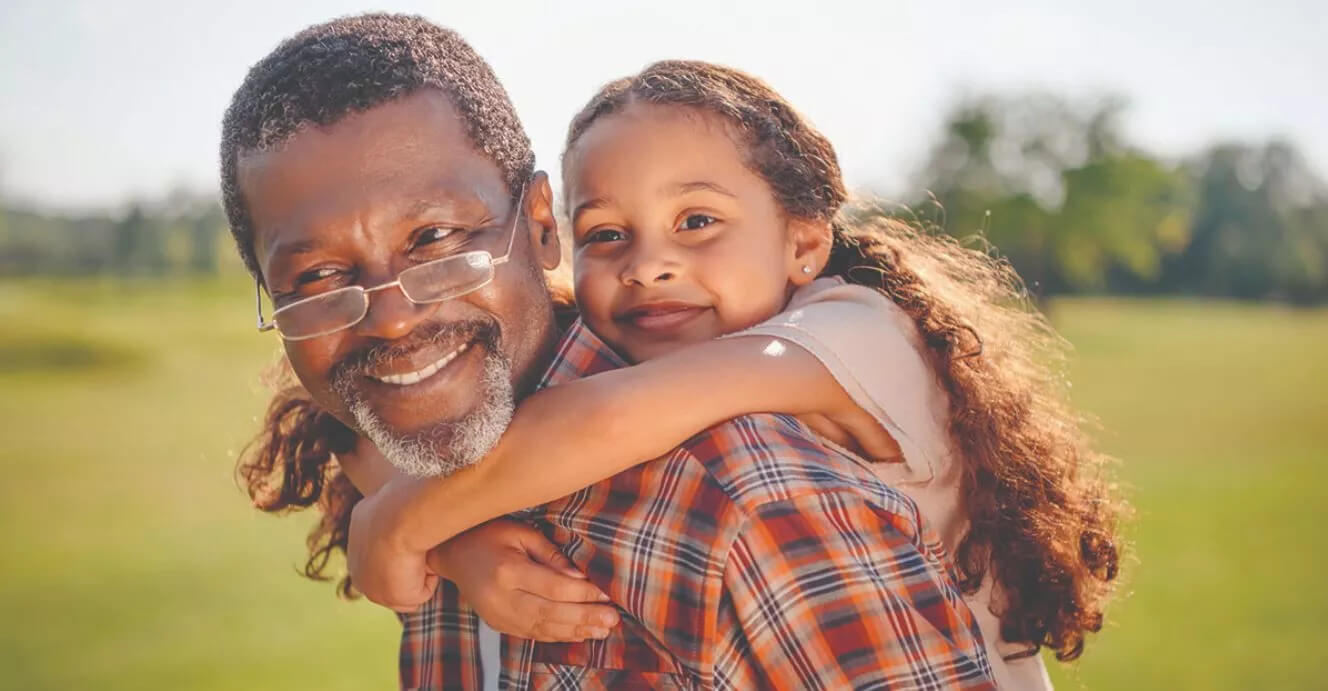 Un homme portant une chemise à carreaux tient une enfant aux longs cheveux bouclés sur son dos dans un parc verdoyant, sous une lumière ensoleillée et chaleureuse.