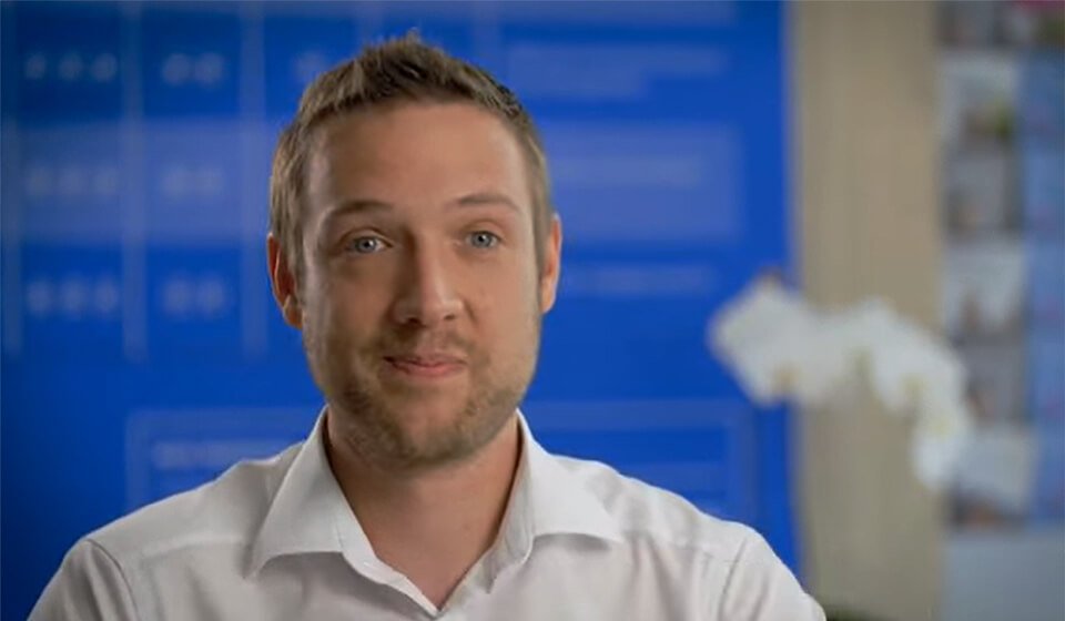 Un homme en chemise blanche, visage flouté, est assis devant un panneau bleu avec des textes flous en arrière-plan, dans un environnement professionnel avec une orchidée blanche sur le côté.