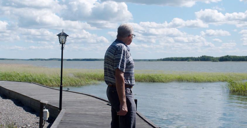 Un homme âgé se tient sur un ponton en bois, regardant un lac entouré de roseaux sous un ciel partiellement nuageux.