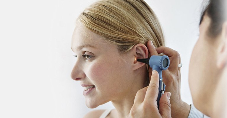 Un audioprothésiste examine l’oreille d’une femme blonde à l’aide d’un otoscope, dans un contexte de centre auditif lumineux, soulignant l’importance des tests auditifs et des soins personnalisés.