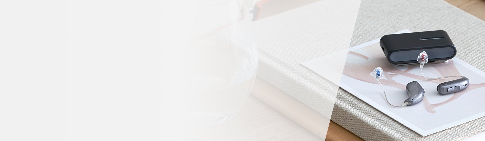 Deux appareils auditifs modernes gris avec embouts colorés (rouge et bleu) reposent près d’un étui de charge noir, posés sur un livre et une table en bois.