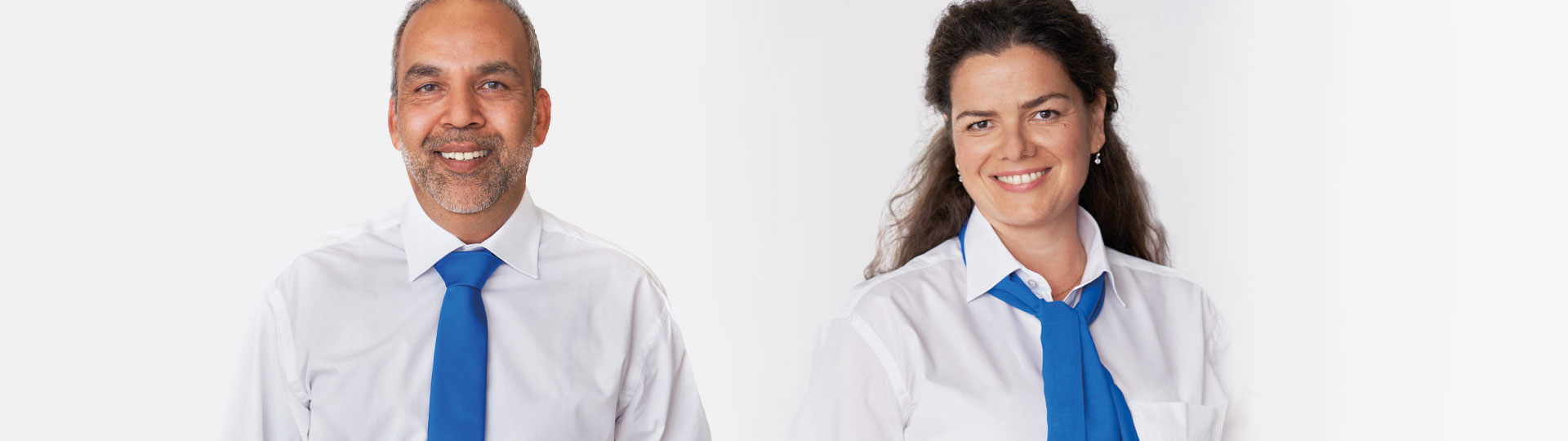 Deux personnes en chemises blanches et cravates bleues, l’une avec des cheveux courts et l’autre avec des cheveux longs et bouclés, posent sur un fond blanc uniforme. Aucune marque ni texte visible.