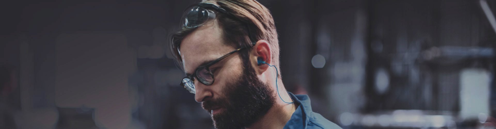 Un homme porte des bouchons d’oreilles bleus reliés par un cordon, dans un environnement industriel sombre. Des lunettes de protection sont positionnées sur sa tête. Aucun texte visible.