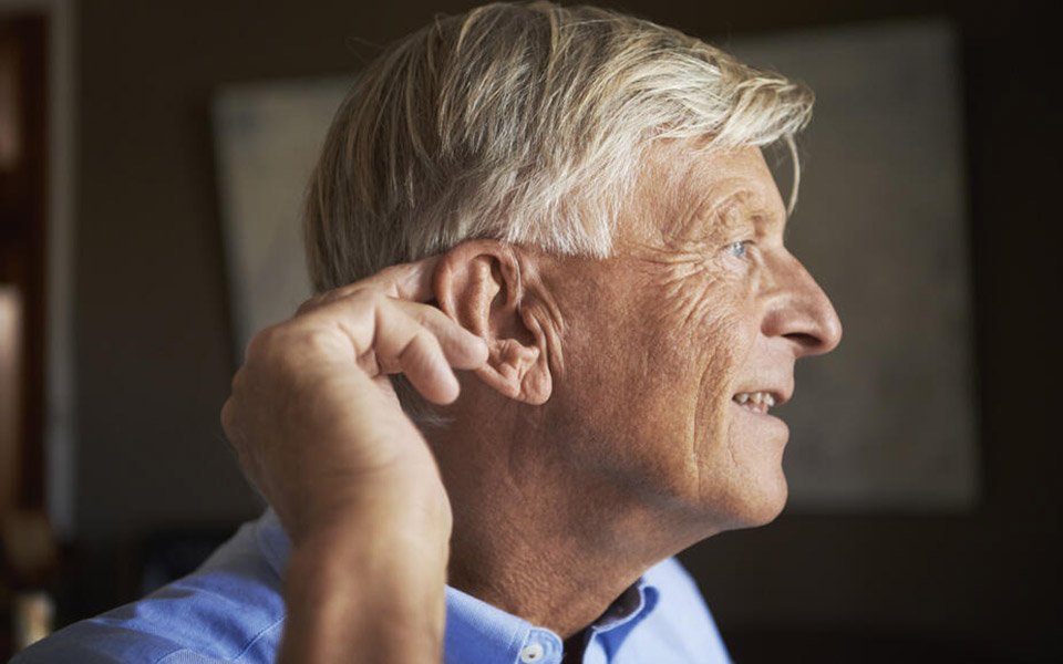 Un homme aux cheveux gris touche son oreille gauche avec la main droite. Il se trouve dans un environnement intérieur aux tons neutres, vêtu d’une chemise bleue. Aucun texte visible.