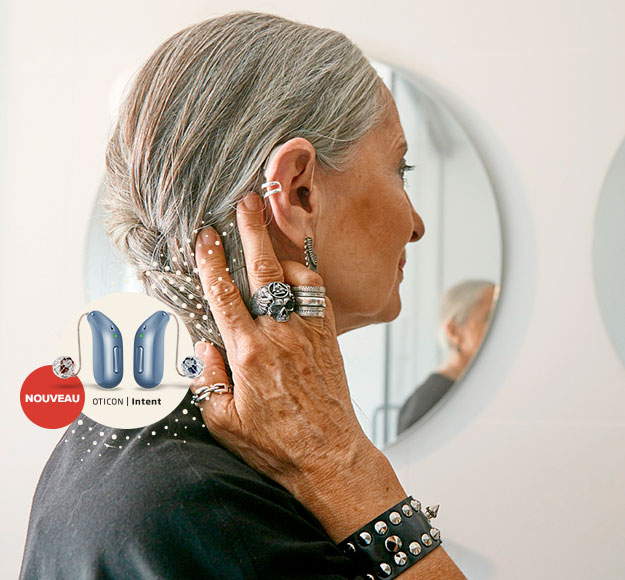Une femme aux cheveux gris ajuste son appareil auditif dans un espace lumineux avec des miroirs circulaires. Texte visible : « NOUVEAU OTICON | Intent », accompagné d’image d’appareils auditifs.