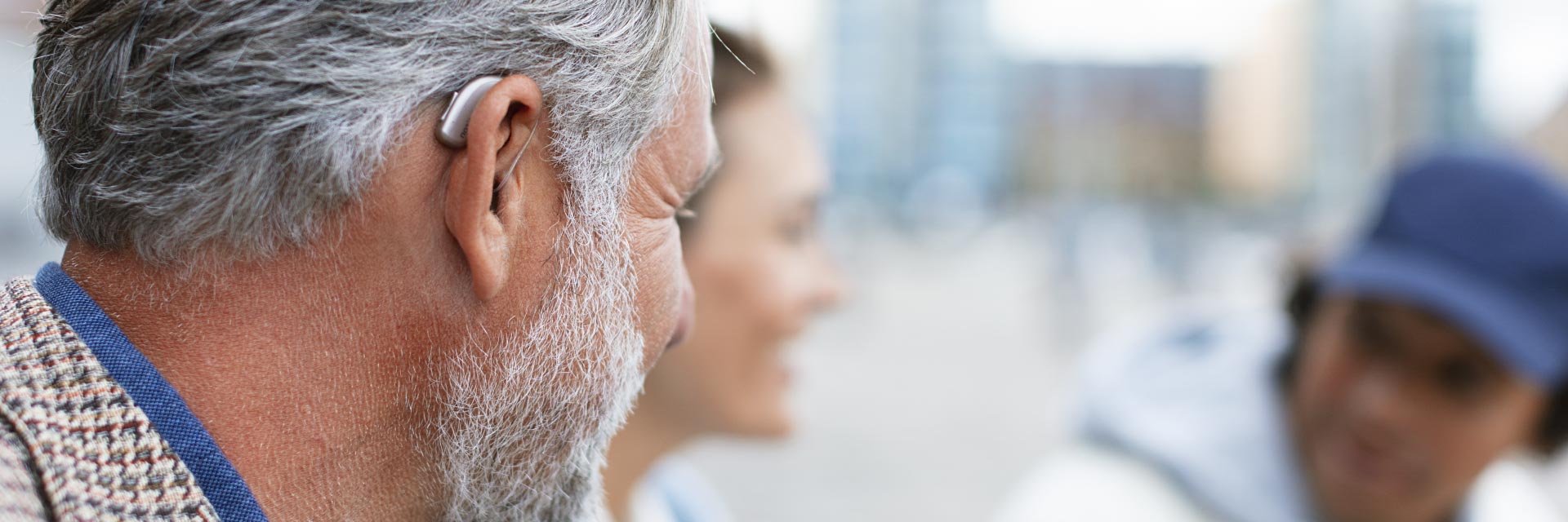 Un homme aux cheveux gris porte un appareil auditif derrière l’oreille gauche. Il est dans un espace urbain flou en arrière-plan, entouré de passants indistincts.