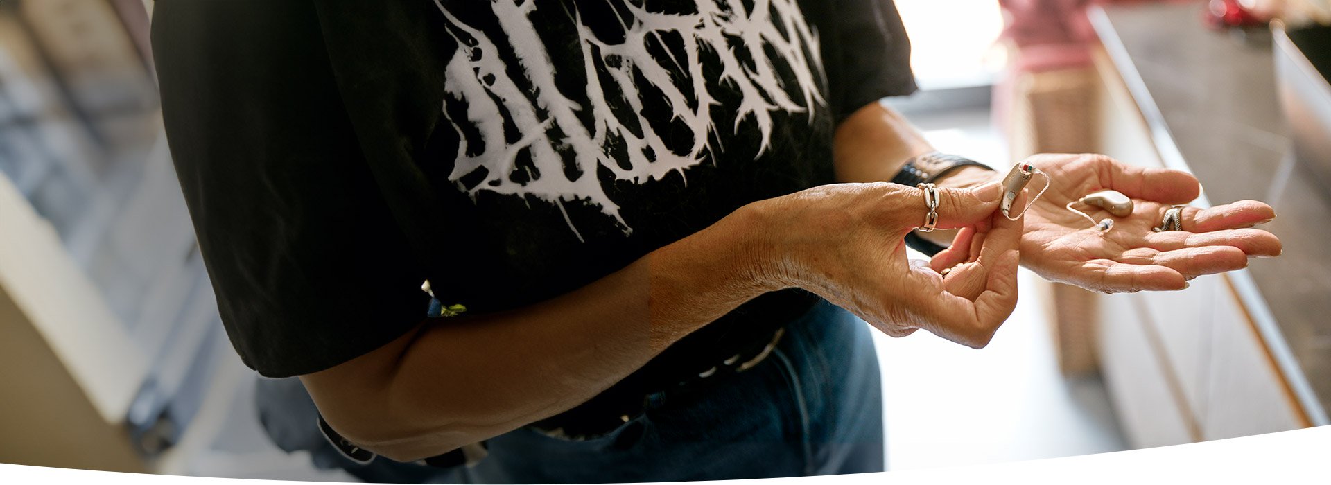 Une personne tient deux appareils auditifs beiges dans ses mains, dans un environnement intérieur lumineux, portant un t-shirt noir avec un motif blanc. Aucun texte ou marque n’est identifiable.