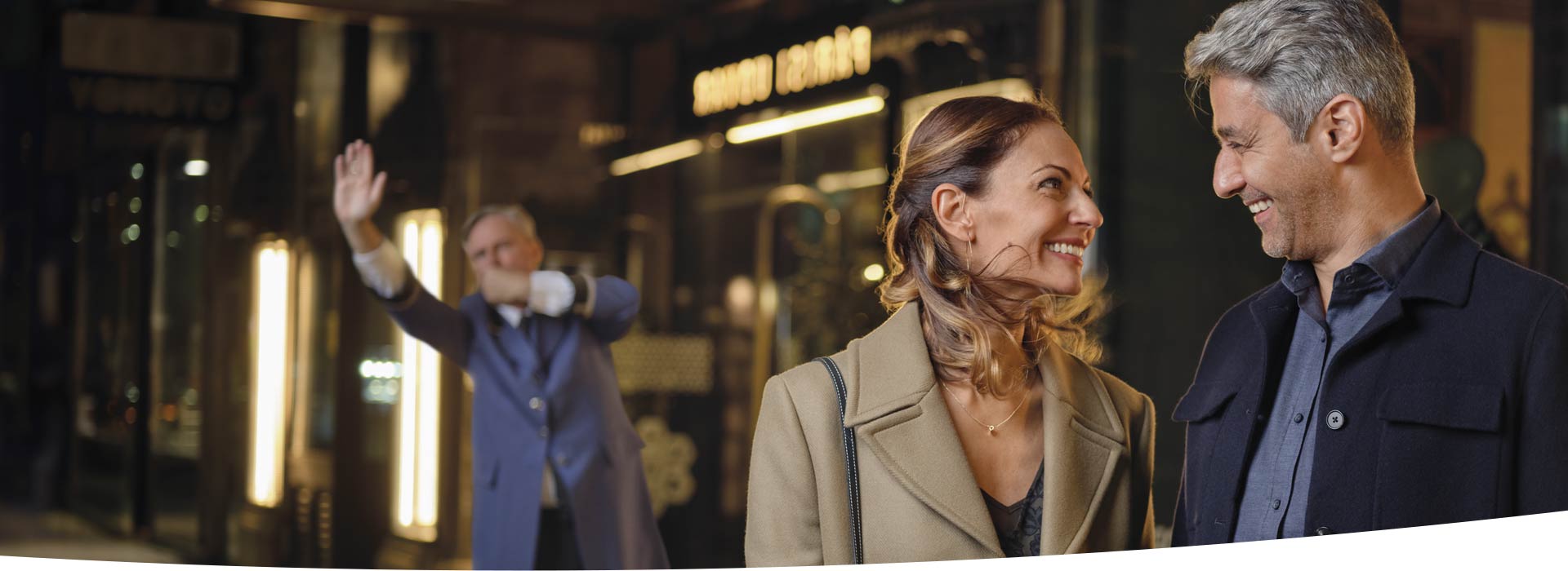 Un couple portant des manteaux élégants marche en soirée devant une enseigne lumineuse. En arrière-plan, un portier en uniforme bleu lève la main près d’une entrée illuminée.