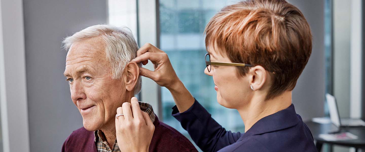Un audioprothésiste ajuste un appareil auditif sur l’oreille d’un patient âgé, dans un cabinet moderne avec des fenêtres lumineuses, représentant un soin professionnel en centre auditif Audika.