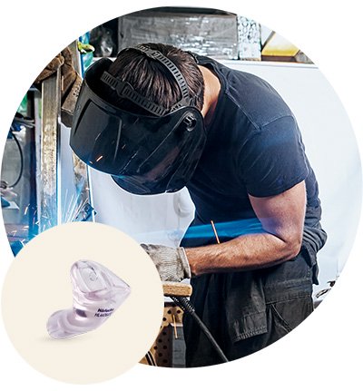 Un ouvrier portant un casque de protection soude du métal, générant des étincelles dans un atelier industriel. En avant-plan, un protecteur d’ouïe moulé Audika est présenté.