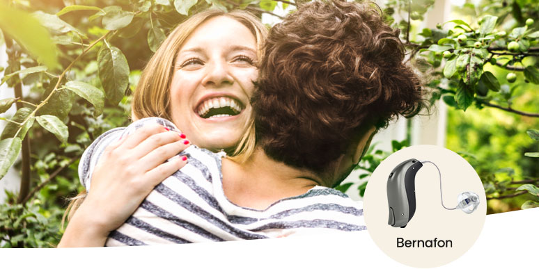 Une personne aux cheveux bouclés enlace un proche dans un jardin verdoyant. En bas à droite, une aide auditive Bernafon est présentée sur un fond beige, mettant en avant l’accompagnement en santé auditive.