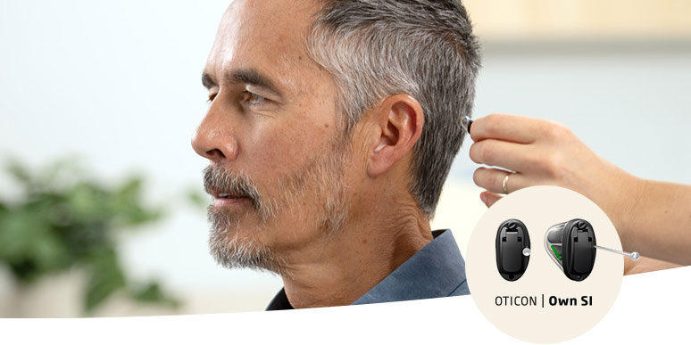 Un audioprothésiste insère un embout auditif dans l’oreille d’un homme aux cheveux gris. À droite, un encart présente deux appareils auditifs noirs avec le texte « OTICON | Own 1 ». En arrière-plan, un environnement lumineux avec des plantes floues complète la scène.