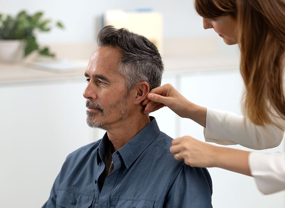 Un audioprothésiste ajuste un appareil auditif sur l’oreille d’un homme aux cheveux gris, dans un environnement lumineux et professionnel, typique d’un centre auditif. Une plante verte est en arrière-plan.