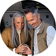 Un couple d’âge mûr, vêtu de vestes décontractées, se tient la main dans un geste affectueux. Le contexte semble être un intérieur chaleureux, probablement un camping-car. Aucun texte visible.