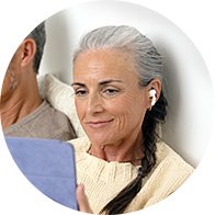 Une femme aux cheveux gris porte un appareil auditif blanc dans son oreille droite, lisant sur une tablette bleue. Un homme est assis à côté, dans un intérieur chaleureux.