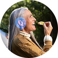 Une femme aux cheveux gris porte un appareil auditif discret derrière l’oreille gauche. Elle tient une chips, semblant apprécier un moment en extérieur dans un cadre verdoyant.