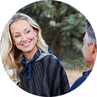 Une femme blonde, vêtue d’un poncho et d’un foulard bleu, discute avec un homme souriant en extérieur, dans un environnement boisé. Elle tient un verre de vin blanc. Aucune mention d’appareil auditif, d’audioprothésiste ou d’Audika visible.