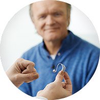 Un audioprothésiste tend un appareil auditif intra-auriculaire à un homme portant un pull bleu dans un contexte de centre auditif lumineux. 