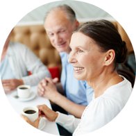 Une femme portant un appareil auditif discute avec deux personnes dans un café lumineux. Des tasses de café sont posées sur la table, créant une ambiance conviviale et chaleureuse.