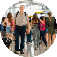 Un homme âgé portant un sac à dos tire une valise à roulettes dans un aéroport animé, entouré de voyageurs en tenue décontractée. Aucun appareil auditif ni texte visible.