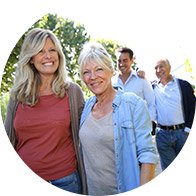 Deux femmes souriantes au premier plan marchent dans un parc verdoyant, suivies de deux hommes également souriants à l’arrière-plan, tous profitant d’un moment convivial en extérieur.