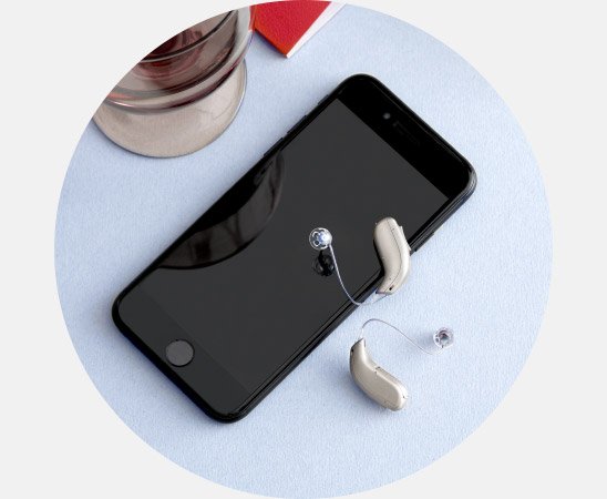 Deux appareils auditifs beige clair sont posés à côté d’un smartphone noir sur une surface bleu clair, à proximité d’un verre et d’un carnet rouge. Aucune marque spécifique n’est identifiable.