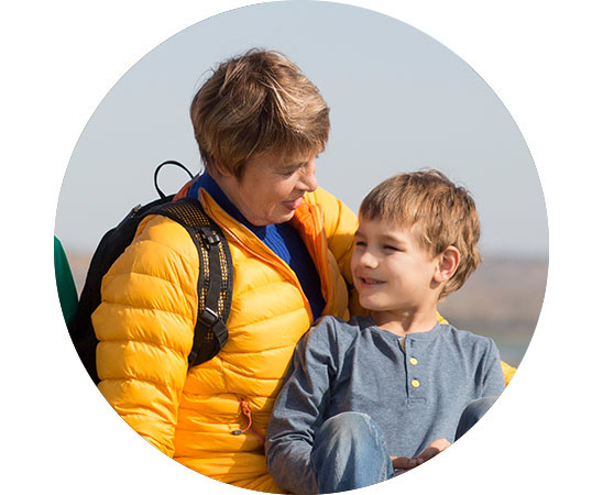 Une personne portant une veste jaune avec un sac à dos noir, tenant un enfant en chemise bleue, est assise à l’extérieur sous un ciel dégagé dans un paysage naturel. Aucun texte visible.