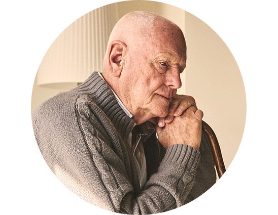 Un homme âgé, portant un pull gris texturé, est appuyé sur une canne en bois dans un intérieur chaleureux, éclairé par une lampe en arrière-plan. Aucune marque d’appareil auditif identifiable.