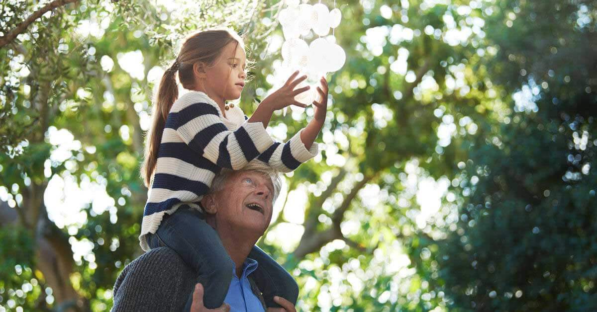 Nonno porta la nipote sulle spalle in un giardino soleggiato.