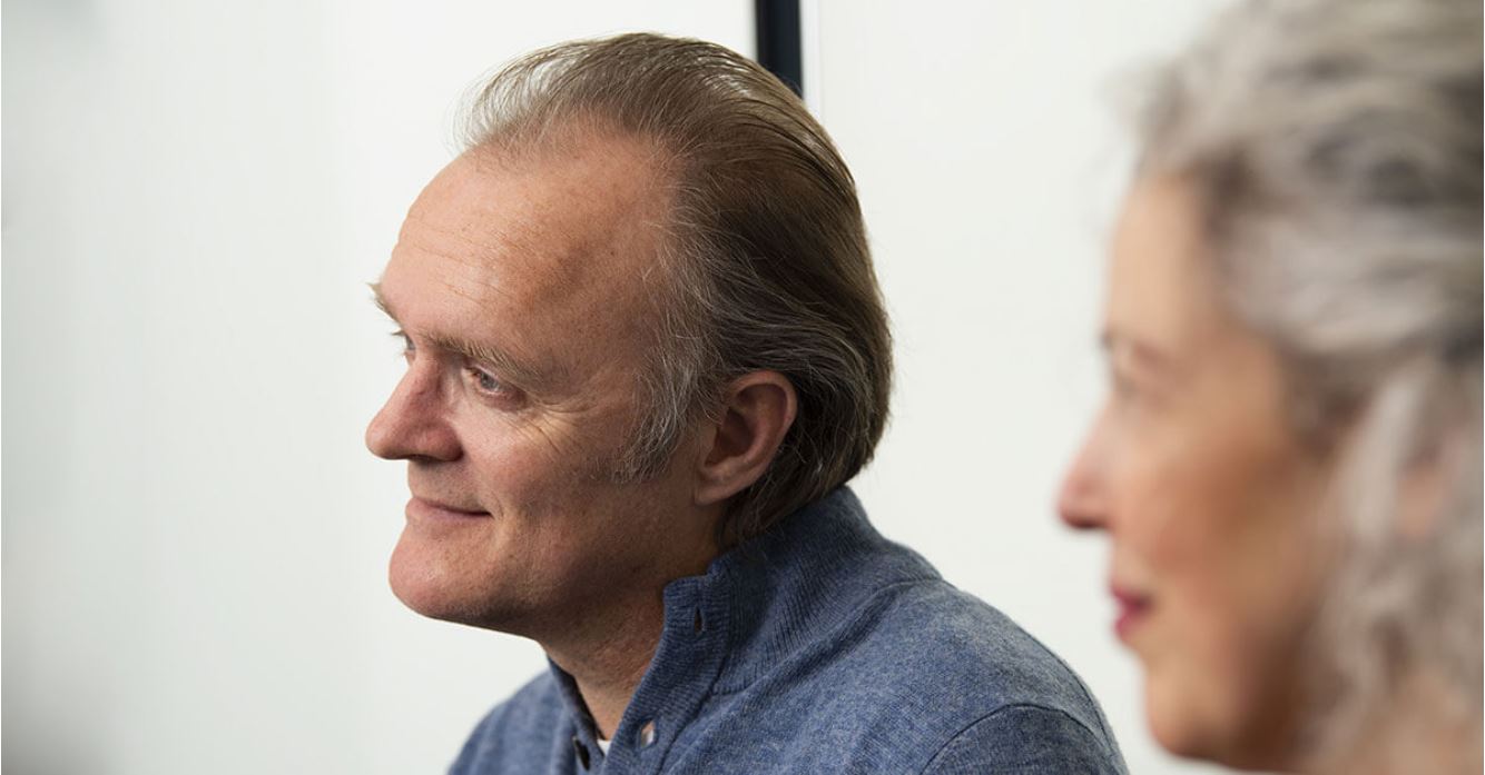 Un uomo con capelli castani e camicia blu è seduto in un ambiente luminoso; sembra essere in una conversazione, forse in una clinica per l'udito Audika. Nessun marchio visibile.
