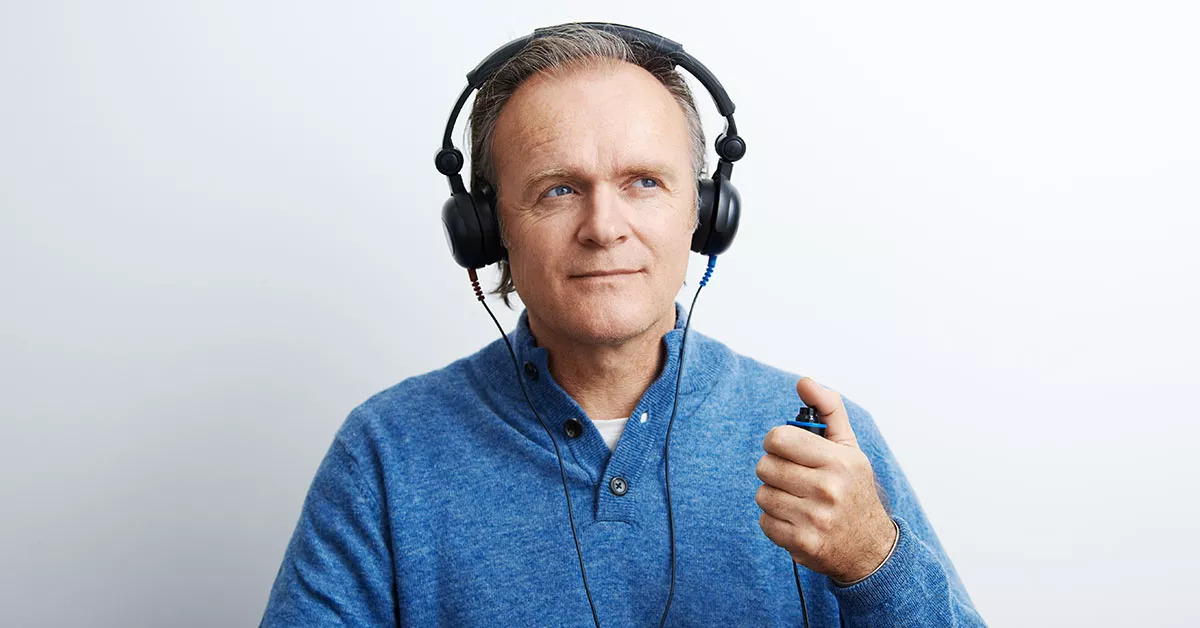 Un uomo indossa cuffie per test dell’udito e tiene un pulsante in mano, indicando una valutazione audiologica in corso. Sfondo bianco, contesto tipico di una clinica dell’udito.
