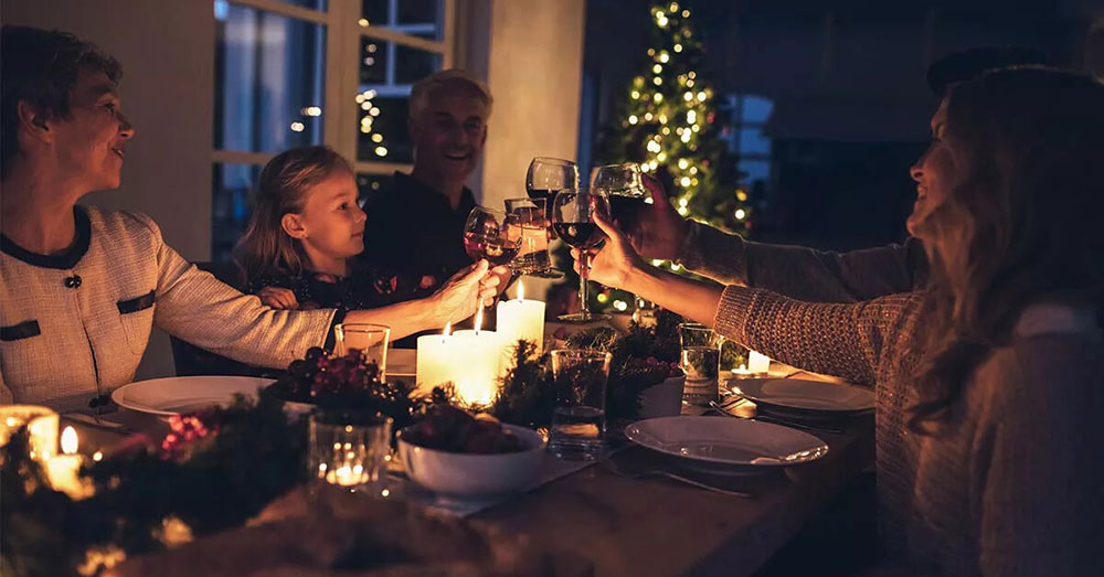 Un gruppo di persone brinda con calici di vino intorno a una tavola apparecchiata, decorata con candele e ornamenti natalizi, in un ambiente interno con albero di Natale illuminato sullo sfondo.