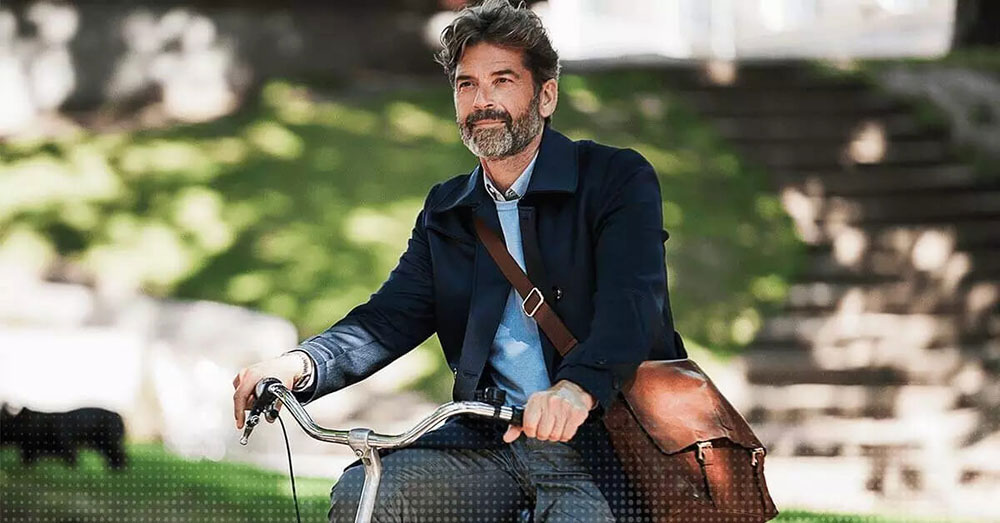 Uomo maturo in bicicletta con una borsa a tracolla, seduto all’aperto in un ambiente verde.