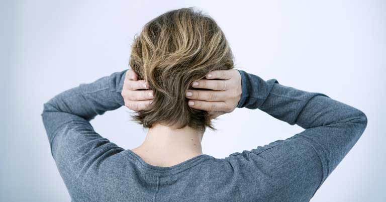Una persona con capelli corti castano chiaro preme entrambe le mani sulle orecchie. Indossa una maglia grigia e si trova in un ambiente neutro con sfondo bianco.